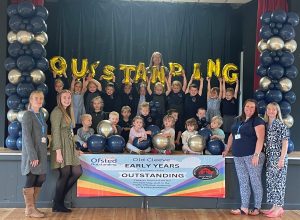 Group of children and staff holding a banner saying Early Years Outstanding, and balloons saying OUTSTANDING.